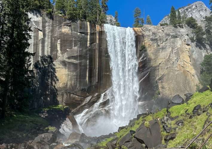 A view of Vernal Falls in Yosemite National Park with the water falling over the granite rocks in the Merced River. Green vegetation can be seen both above the falls and on the land either side of the river into which the water flows.