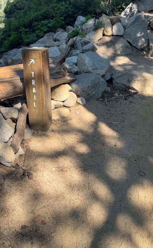 A small wooden sign located on the edge of the Echo Lakes Trail as it winds its way around Lower Echo Lake.. The sign has an arrow and the word "trail" in white lettering.