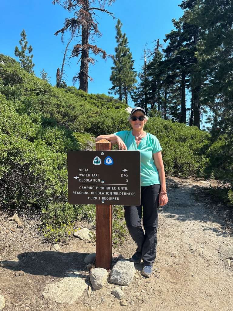 The author of this blog post is seen standing next to a trail sign on the Echo Lakes Trail. She is wearing a baseball cap and sunglasses, a mint green shirt, black hiking trousers and hiking shoes. She is looking into the camera. 