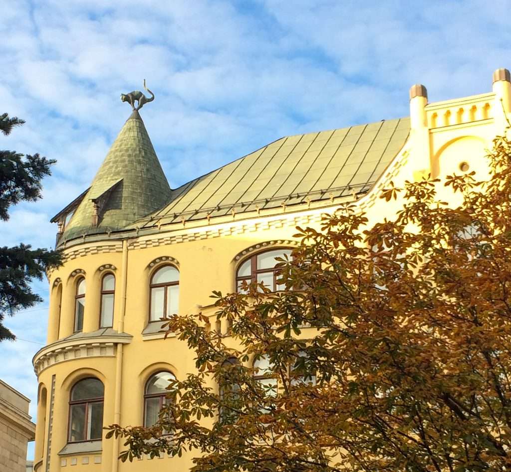 A view of the black cat that sits on top of the Cat House in Riga. The Cat House is a large building and has a distinctive yellow colour and turret. A tree with brownish coloured foliage is visible in the foreground.