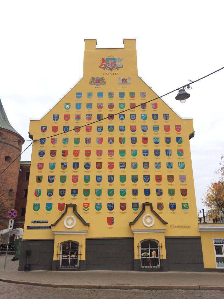 Image shows a close up of a yellow coloured building located close to the Powder Tower in Riga. The building is known as Jacobs Barracks. Multiple coats of arms decorate the external elevation of part of the building.