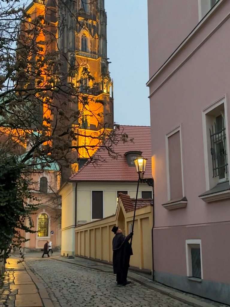 Lamplighter of Wroclaw lighting a gas lamp early one evening with the Cathedral in the background and glowing beneath spotlights.