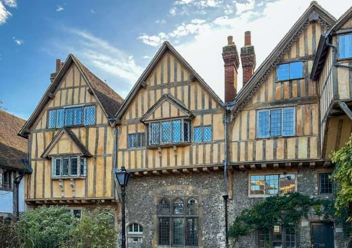 Three half-timbered buildings stand in a row in Cheney Court in Winchester. They are A-framed in shape and each one has several windows. Two chimneys are visible.