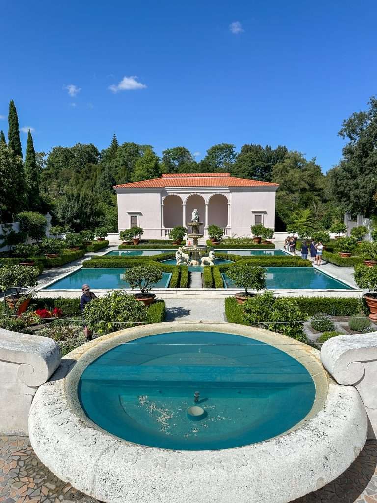 The Italian Renaissance Garden at Hamilton Gardens is characterised by a central pool with water divided into 4 squares surrounded by low level planting. In the middle of the pool is an Italian marble statue. In the foreground is a small circular pool and in the background is a plain cream Italian structure with russet red roof and 3 arches forming a portico.