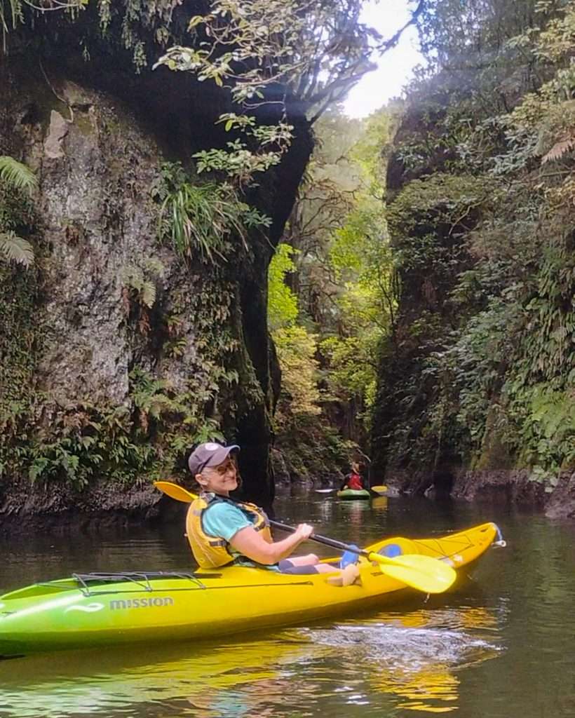 The author of this blog posts is in a kayak on the water and smiling into the camera. The kayak and paddle are yellow with some green colouring toward the rear of the kayak. The kayaker is wearing a blue cap, blue shorts, mint green t-shirt, blue water shoes and yellow/black life jacket.