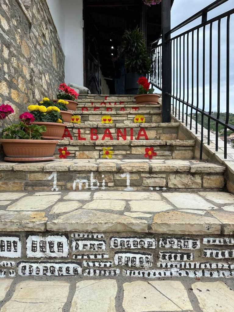 Stone steps lead up to a restaurant in the city of Berat. Red and yellow flowers in pots line up along the left side of the stairs whilst on the right is an iron railing. The steps feature the word Albania written in red and a black and white picture of the Ottoman houses that line the hills in Berat.