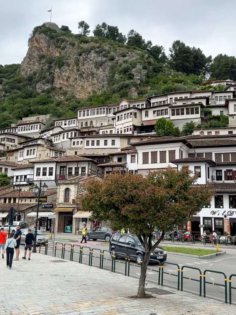 A view of the city of Berat with the pavement and road visible in the image. Pedestrians can be seen walking on the pavements and a black car drives past. A tree on the pavement partially blocks a view of the car. The backdrop to the pavement and road are the houses of Berat with all their windows and high above them the viewing point within the castle and the flagpole can be seen.
