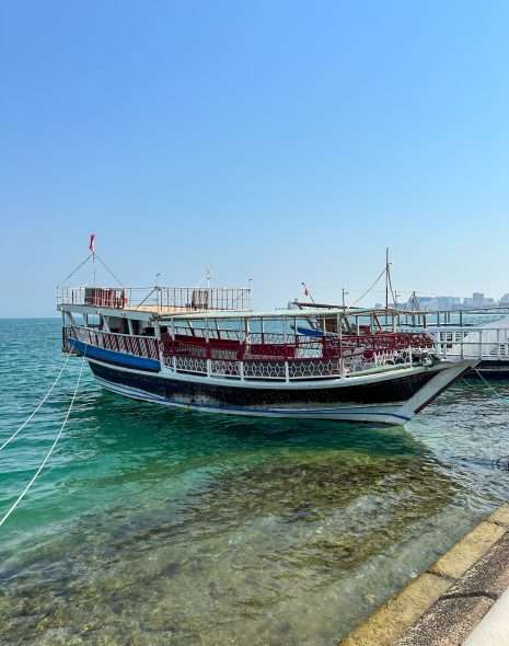 A white and blue coloured wooden sailing vessel known as a dhow sits in Doha Bay. White ropes attaching it to the shore are visible.