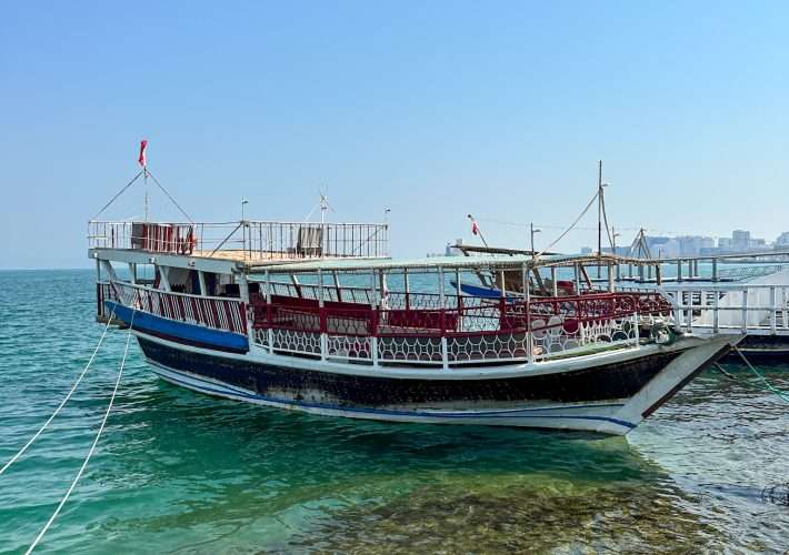 A white and blue coloured wooden sailing vessel known as a dhow sits in Doha Bay. White ropes attaching it to the shore are visible.
