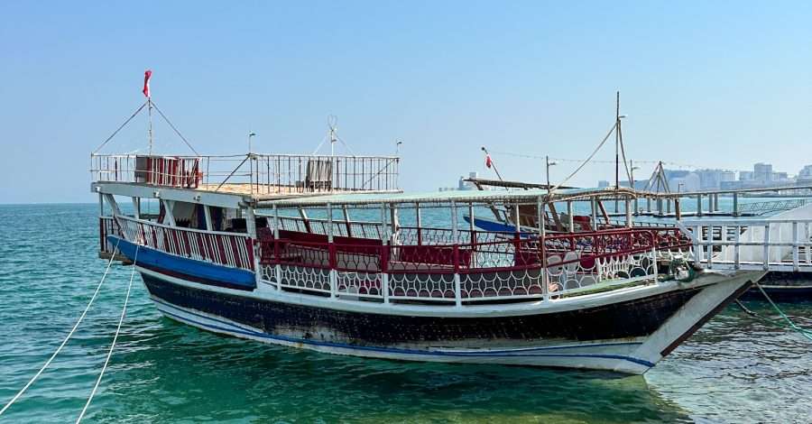 A white and blue coloured wooden sailing vessel known as a dhow sits in Doha Bay. White ropes attaching it to the shore are visible.