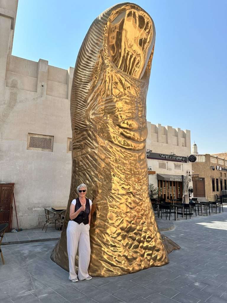a large golden statute of a thumb located in the Souq Waqif in Doha. Sarah, the author of this blog post, is pictured next to the thumb which towers over her.