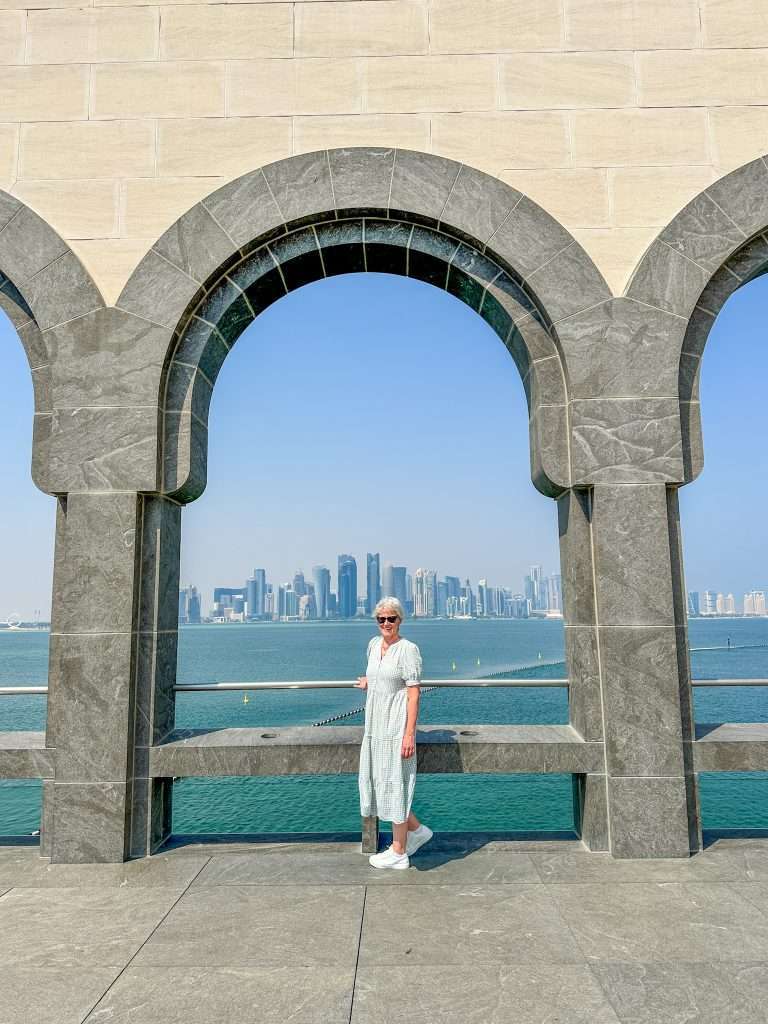 The author of this blog post poses in front of archways on the terrace at the Museum of Islamic Art in Doha with the city skyline visible across Doha Bay in the background.