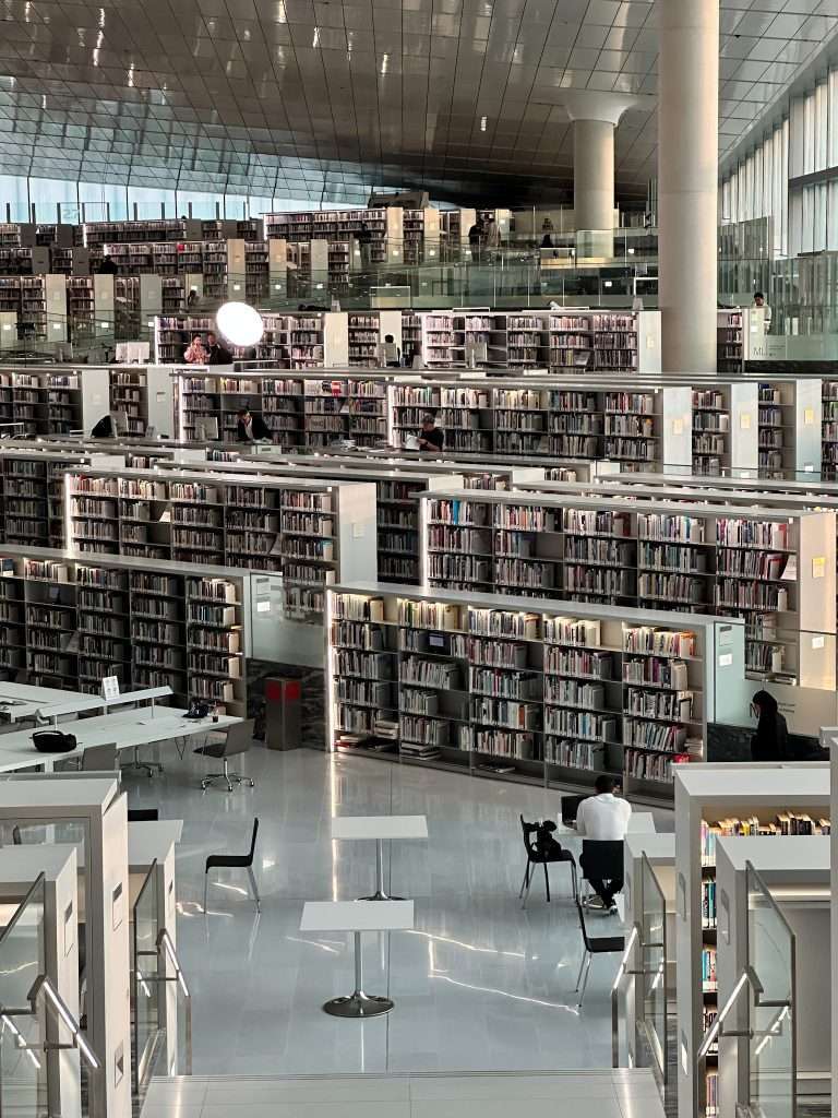 An internal view of the National Library of Qatar shows many white shelves of books reaching back into one corner of the diamond shaped building. A person can be seen sitting at one of the desks whilst other visitors are dotted about the interior.