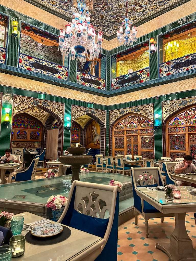 The main dining room at Parisa restaurant with ornate ceilings and chandeliers, a fountain in the centre and dining booths lining the main restaurant. Islamic inspired shapes are visible in the archways and doors within the dining room.