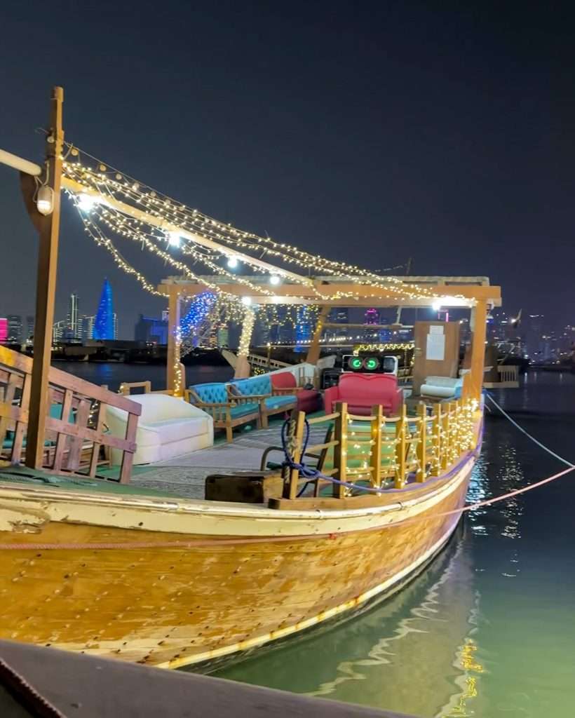A wooden dhow is moored alongside the Corniche in Doha. The image is taken in the evening with lights strung along the boat shining in the dark. Several seats coloured blue and red are visible along the side of the dhow, ready for passengers.